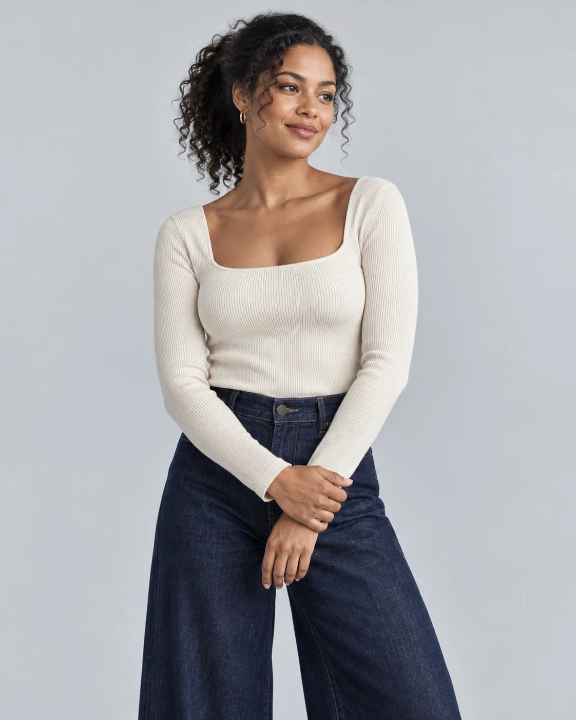 Model wearing an ivory knit top and dark denim against a grey studio backdrop